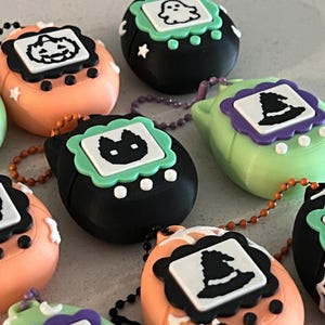 May include: A collection of Halloween-themed keychains in various colors, including orange, black, and green. Each keychain features a different Halloween icon, such as a pumpkin, ghost, cat, witch hat, and skull. The keychains are attached to beaded chains.