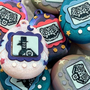 May include: A collection of colorful, handheld digital pet toys. Each toy features a small screen displaying a pixelated graphic, surrounded by a decorative frame. The toys are in various colors, including blue, pink, and purple, with star and moon accents.