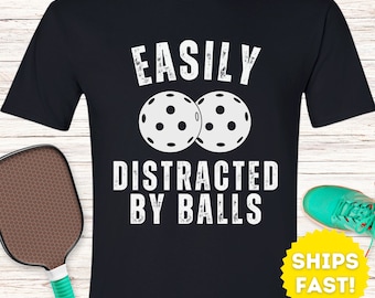 May include: Black t-shirt with white text that reads "Easily Distracted By Balls" and a graphic of two white pickleball paddles.