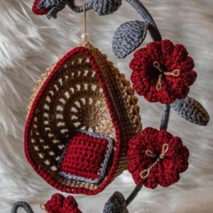 May include: A crocheted hanging chair with a red cushion, suspended from a gray crocheted branch with red flowers. The text "Cozy Cocoon" is visible in the top left corner of the image.