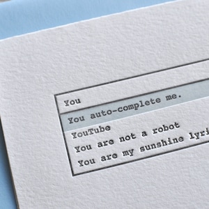 May include: A white card with a gray search bar and text that reads "You auto-complete me."  The search bar has a magnifying glass icon. Below the search bar are three lines of text that read "YouTube", "You are not a robot", and "You are my sunshine lyrics".