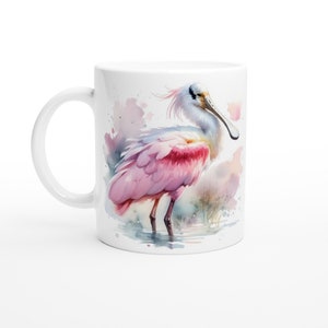 May include: White ceramic mug with a watercolor illustration of a pink roseate spoonbill standing in a shallow body of water. The spoonbill has a long, thin beak and pink feathers.