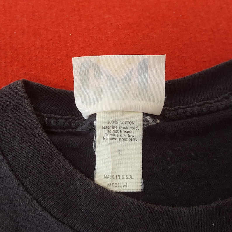 May include: Close-up of a black t-shirt with a white tag. The tag has the letters "CVI" at the top and includes washing instructions and the text "MADE IN U.S.A. MEDIUM". The shirt is made of 100% cotton.
