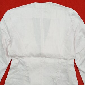 May include: A white long-sleeved blazer with a V-neck design. The back of the blazer is visible, showcasing the tailored seams and the fabric's texture. The garment is made of a lightweight material, suitable for layering.