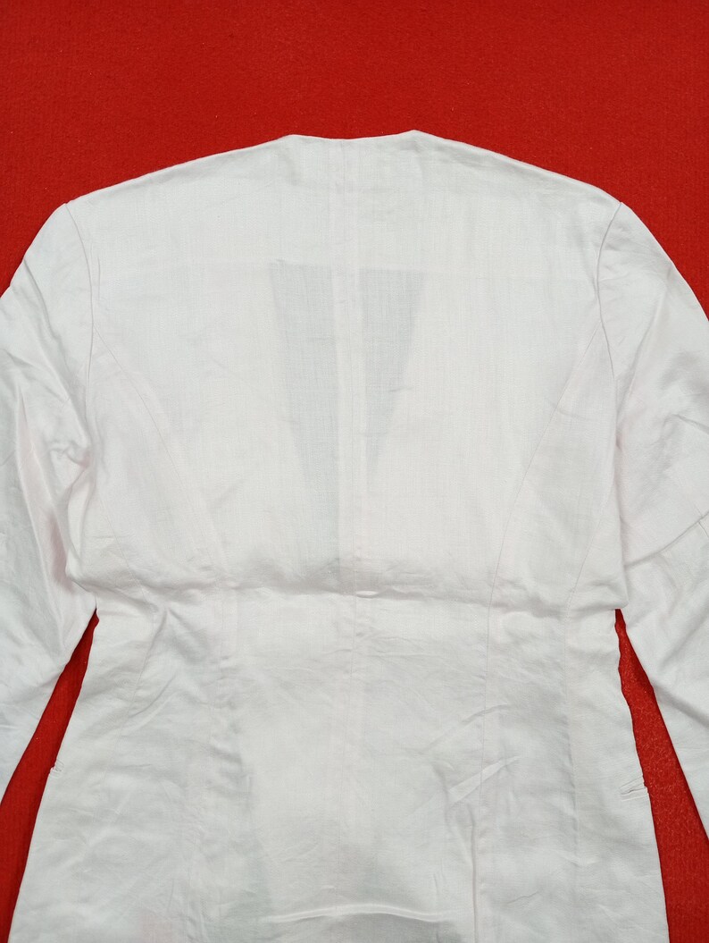 May include: A white long-sleeved blazer with a V-neck design. The back of the blazer is visible, showcasing the tailored seams and the fabric's texture. The garment is made of a lightweight material, suitable for layering.