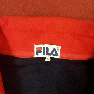 May include: A red fleece jacket with a black Fila logo tag. The tag has the word "FILA" in white letters and a size "L" tag.