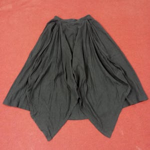 May include: A black skirt with a unique asymmetrical design. The skirt features a high waist and a flowing, draped silhouette. The fabric appears to be lightweight and has a soft texture. The skirt is spread out on a red surface.