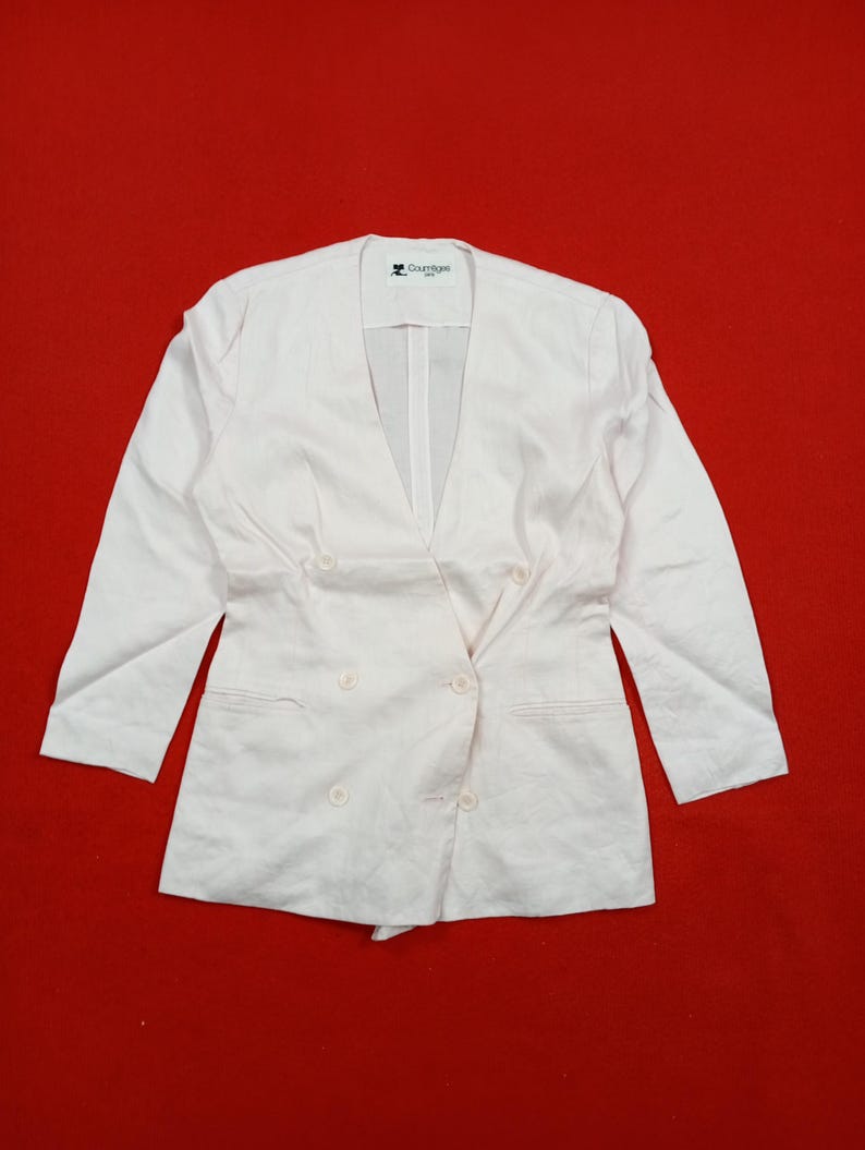 May include: A light pink double-breasted blazer with a V-neckline and three-quarter sleeves. The blazer has two rows of buttons and a tailored fit. The Courreges logo is visible on the upper left side of the blazer.