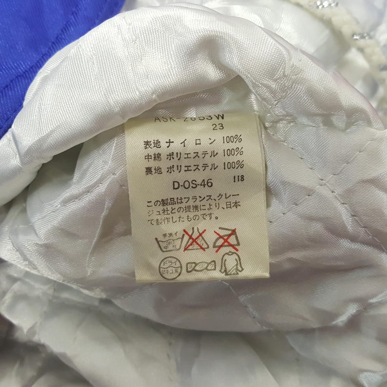 Puede incluir: Primer plano de una etiqueta de ropa con texto japon&eacute;s y s&iacute;mbolos de cuidado. La etiqueta incluye el texto "ASK-2053W 23" y "D-OS-46 118". La etiqueta est&aacute; sobre una tela acolchada blanca, con tela azul visible en el fondo.