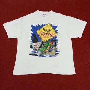 May include: White t-shirt with a graphic print of a cartoon frog in a boat. The graphic also includes a yellow sign with the text "HIGH WATER MARKS" and a martini glass with the text "TOYOTA WE'RE WITH YOU ALWAYS".