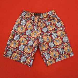 May include: Red shorts featuring a repeating pattern of orange pumpkins and blue flowers. The shorts have a black waistband and a small pocket. Made from lightweight fabric, these shorts are ideal for warm weather.