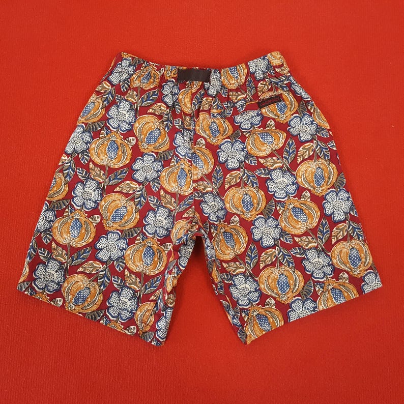 May include: Red shorts featuring a repeating pattern of orange pumpkins and blue flowers. The shorts have a black waistband and a small pocket. Made from lightweight fabric, these shorts are ideal for warm weather.