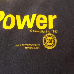May include: Close-up of a black fabric with yellow text that reads "Power". Below the text is the copyright information for Caterpillar Inc. 1993. The image also includes the text "S.O.S. ENTERPRISES LTD. MARION, IOWA" and a CAT logo.