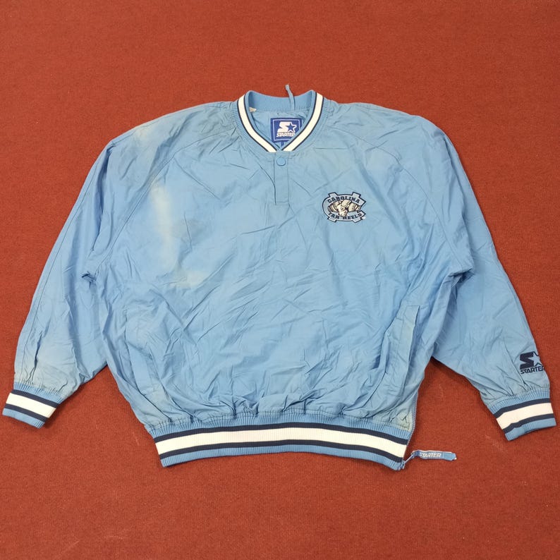 Pu&ograve; includere: Giacca a vento blu chiaro con scollo a V e maniche lunghe. Presenta un logo Carolina Tar Heels sul petto e un logo Starter sulla manica. I polsini e l'orlo hanno strisce bianche e blu navy.