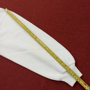 May include: A white long-sleeved sweatshirt sleeve with a yellow measuring tape laid across it. The measuring tape shows measurements in inches, with the sleeve extending to approximately 24 inches.