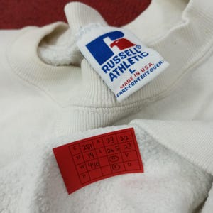 May include: Close-up of a white Russell Athletic sweatshirt. The tag reads "Russell Athletic L MADE IN USA CARE-CONTENT OVER". A red tag with handwritten numbers is also visible.