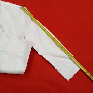 May include: A white long-sleeved shirt with a measuring tape laid across the sleeve. The shirt is on a red background. The measuring tape shows measurements in inches.