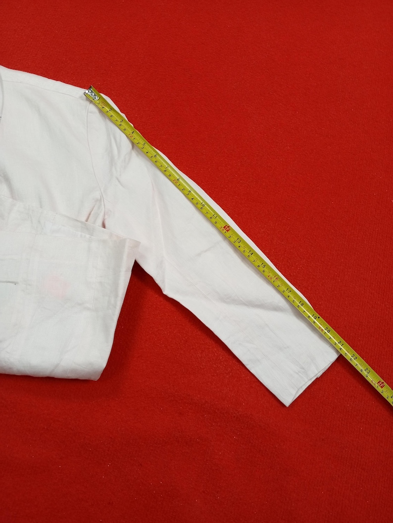 May include: A white long-sleeved shirt with a measuring tape laid across the sleeve. The shirt is on a red background. The measuring tape shows measurements in inches.
