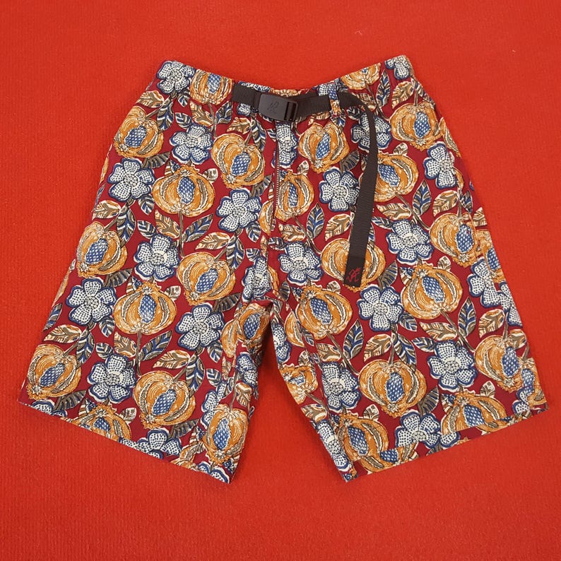 May include: Shorts featuring a colourful floral design in orange, blue, and white on a burgundy backdrop. They have a black belt and buckle. The shorts are made of a lightweight material.