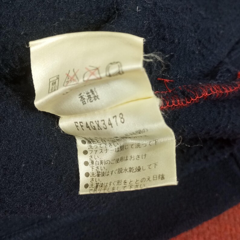 May include: A white tag with black text and symbols. The tag has the text "FF4GX3478" and instructions for washing and caring for the garment in Japanese. The tag is attached to a dark blue garment.