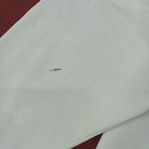 May include: Close-up of a light gray fabric with a small dark stain and a yellow stain. The fabric appears to be part of a garment, with a red fabric visible in the corner.