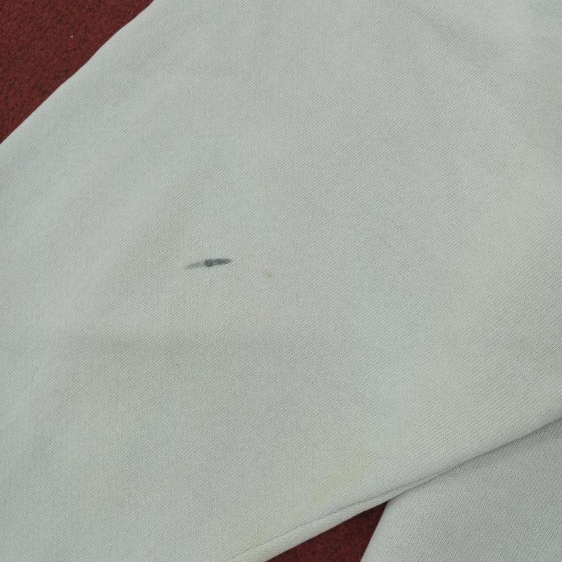 May include: Close-up of a light gray fabric with a small dark stain and a yellow stain. The fabric appears to be part of a garment, with a red fabric visible in the corner.