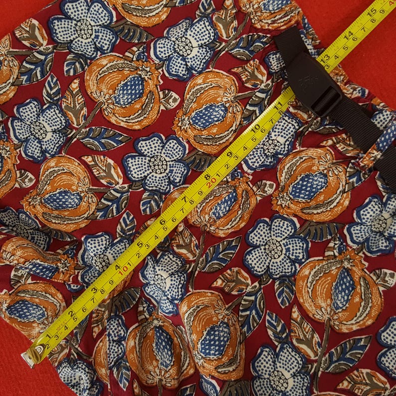 May include: Red fabric with a floral design of orange, blue, and white flowers and fruit. A yellow measuring tape indicates a length of approximately 38 cm. A black strap is also visible.