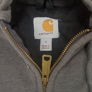 May include: A gray Carhartt zip-up hoodie with a black hood and a gold zipper. The tag says "Carhartt" and "Made in Mexico".