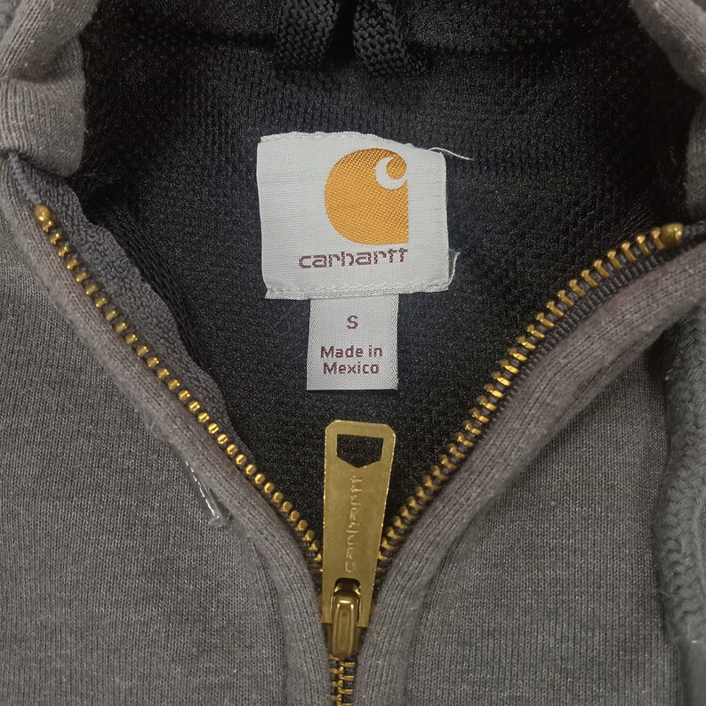 May include: A gray Carhartt zip-up hoodie with a black hood and a gold zipper. The tag says "Carhartt" and "Made in Mexico".