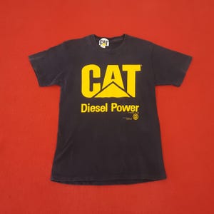 May include: Black t-shirt with a yellow "CAT" logo and "Diesel Power" text. The shirt has a crew neck and short sleeves. A small "CAT" logo is on the collar.
