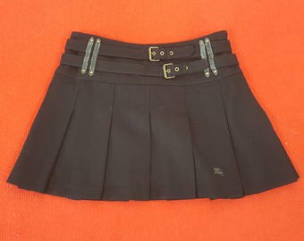 May include: A black pleated mini skirt with two buckled belts and leather accents. The skirt has a small logo near the hem. The skirt is made of a dark fabric.