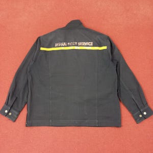 Puede incluir: Chaqueta azul marino oscuro con mangas largas y puños abotonados. La parte trasera de la chaqueta presenta el texto "JAPAN POST SERVICE" en letras naranjas sobre una franja amarilla. La chaqueta tiene cierre de cremallera y cuello mandarín.