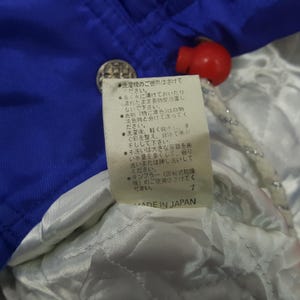 Puede incluir: Primer plano de una chaqueta azul con forro blanco y un cierre rojo. Una etiqueta con texto japon&eacute;s es visible, junto con un bot&oacute;n plateado. La chaqueta est&aacute; hecha en Jap&oacute;n.