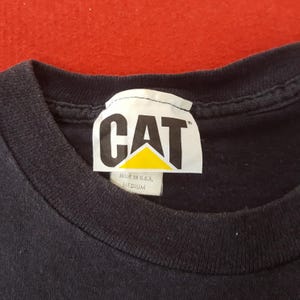 May include: Close-up of a black t-shirt with a white tag. The tag features the word "CAT" in bold, black letters above a yellow triangle. The tag also reads "MADE IN U.S.A." and "MEDIUM".
