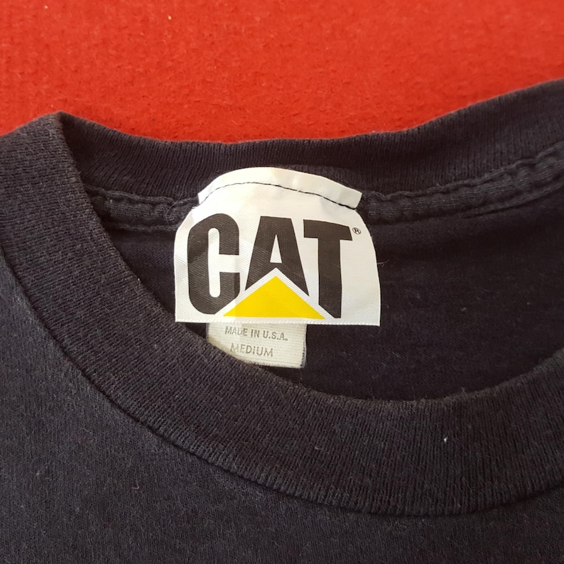 May include: Close-up of a black t-shirt with a white tag. The tag features the word "CAT" in bold, black letters above a yellow triangle. The tag also reads "MADE IN U.S.A." and "MEDIUM".