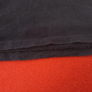 May include: Close-up of a dark navy blue fabric with a raw, unfinished edge. The fabric is layered over a vibrant red surface, creating a contrast in color and texture. The image highlights the details of the textile.