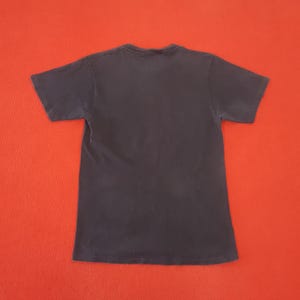 May include: A dark navy blue short-sleeved t-shirt, viewed from the back, is centered against a bright red background. The shirt's fabric appears soft and slightly worn, with a simple crew neck design.