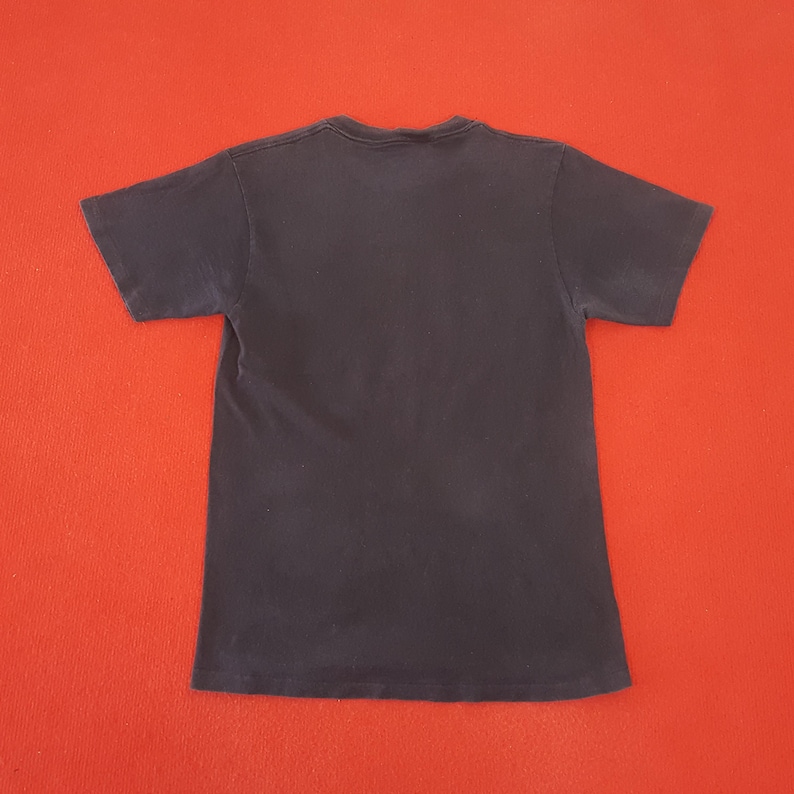 May include: A dark navy blue short-sleeved t-shirt, viewed from the back, is centered against a bright red background. The shirt's fabric appears soft and slightly worn, with a simple crew neck design.