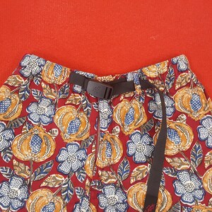 May include: Close-up of patterned shorts with a dark brown belt. The shorts feature a red background with a repeating pattern of orange gourds, blue and white flowers, and brown leaves. The belt has a black buckle.