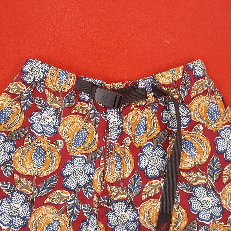 May include: Close-up of patterned shorts with a dark brown belt. The shorts feature a red background with a repeating pattern of orange gourds, blue and white flowers, and brown leaves. The belt has a black buckle.