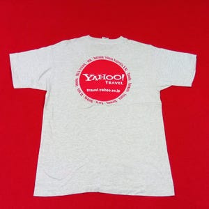 Könnte beinhalten: Hellgraues T-Shirt mit einem großen roten Kreis-Grafik auf der Rückseite. Die Grafik zeigt das Yahoo! Travel Logo und den Text "travel.yahoo.co.jp". Das T-Shirt liegt auf rotem Hintergrund.