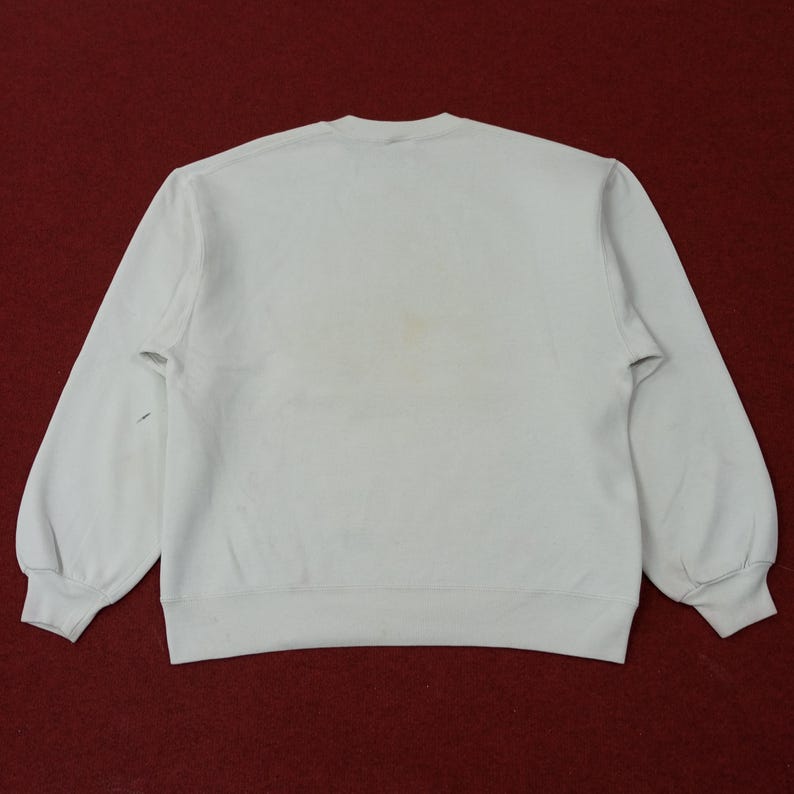 May include: A light-colored sweatshirt with long sleeves, viewed from the back. The garment has a crew neck and ribbed cuffs and hem. There are some stains on the front of the sweatshirt. The background is a dark red.