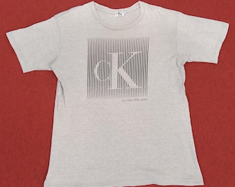 May include: A gray short-sleeved t-shirt with a black and white graphic of the letters "CK" in a striped pattern. The text "CK Calvin Klein Jeans" is printed below the graphic.