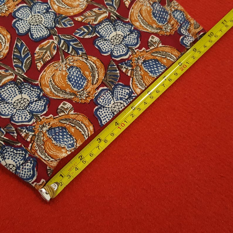 May include: A square fabric featuring a repeating pattern of orange and blue floral and pumpkin designs on a burgundy background. A yellow tape measure shows the fabric is approximately 25 cm long.