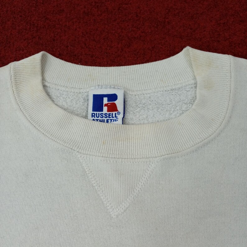 May include: Close-up of a cream-colored Russell Athletic sweatshirt. The crew neck and the bottom of the sweatshirt have a stitched design. The Russell Athletic logo is visible on the tag, featuring a blue "R" and the brand name.