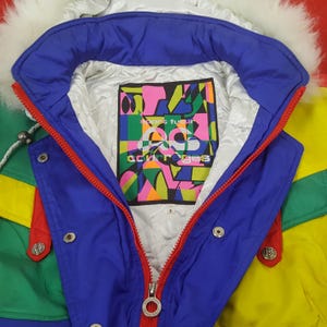 Puede incluir: Chaqueta de esqu&iacute; vintage colorida con cuello de piel sint&eacute;tica blanca. La chaqueta presenta un cuello azul, cremallera roja y detalles en verde y amarillo. El forro interior tiene un gr&aacute;fico colorido con el texto "acurr&egrave;ges".