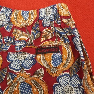 May include: Close-up of red shorts with a colourful floral design in orange, blue, and white. The shorts have a small pocket with the brand name "Gramicci" on a black label.