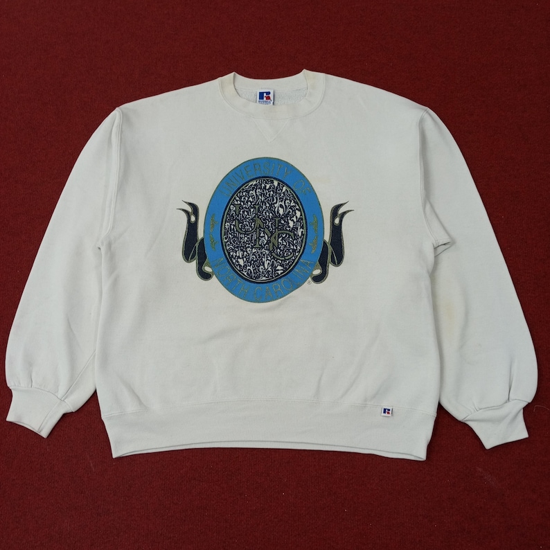 May include: Off-white crew neck sweatshirt with a blue and white circular graphic on the front. The graphic has the word "RIVER" in a blue circle with a decorative design in the center. The sweatshirt has long sleeves and a ribbed hem.