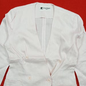 May include: A light pink double-breasted blazer with a V-neckline and four buttons. The blazer has a Courr&egrave;ges Paris label. The fabric appears to be linen or a linen blend. The blazer is on a red background.