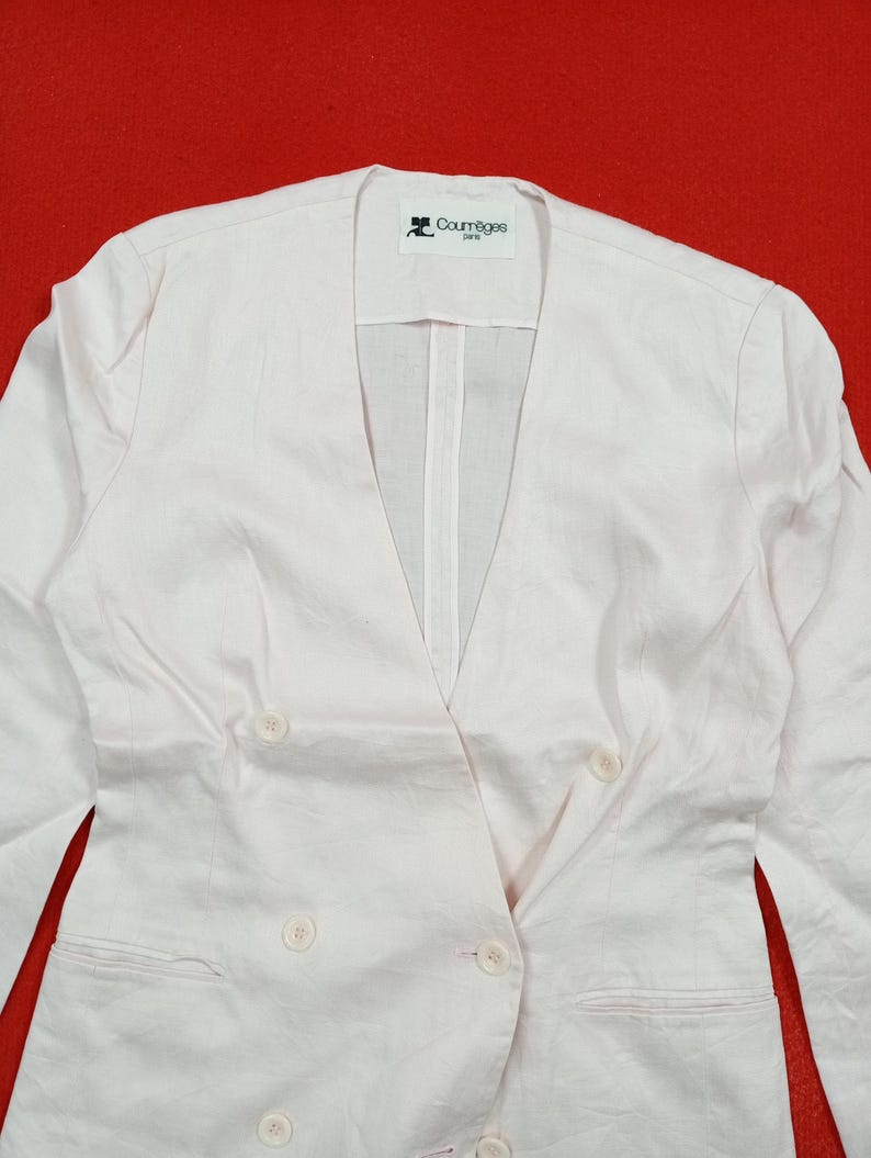 May include: A light pink double-breasted blazer with a V-neckline and four buttons. The blazer has a Courr&egrave;ges Paris label. The fabric appears to be linen or a linen blend. The blazer is on a red background.